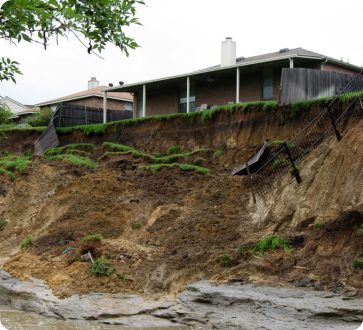 A house sits on a hillside with significant erosion, exposing soil and a damaged fence. Green grass peeks through the dirt.