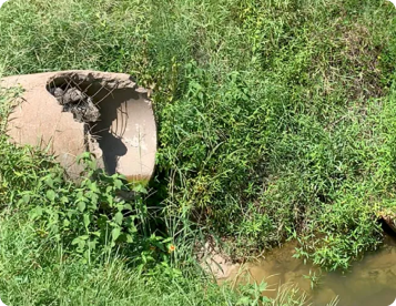 A house sits on a hillside with significant erosion, exposing soil and a damaged fence. Green grass peeks through the dirt.