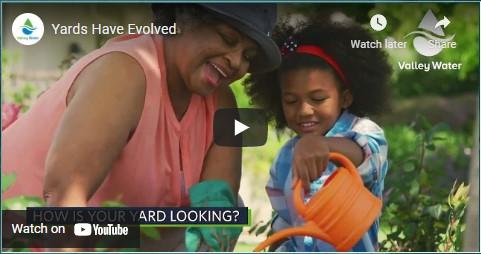 An adult and child smiling as they water plants outdoors.