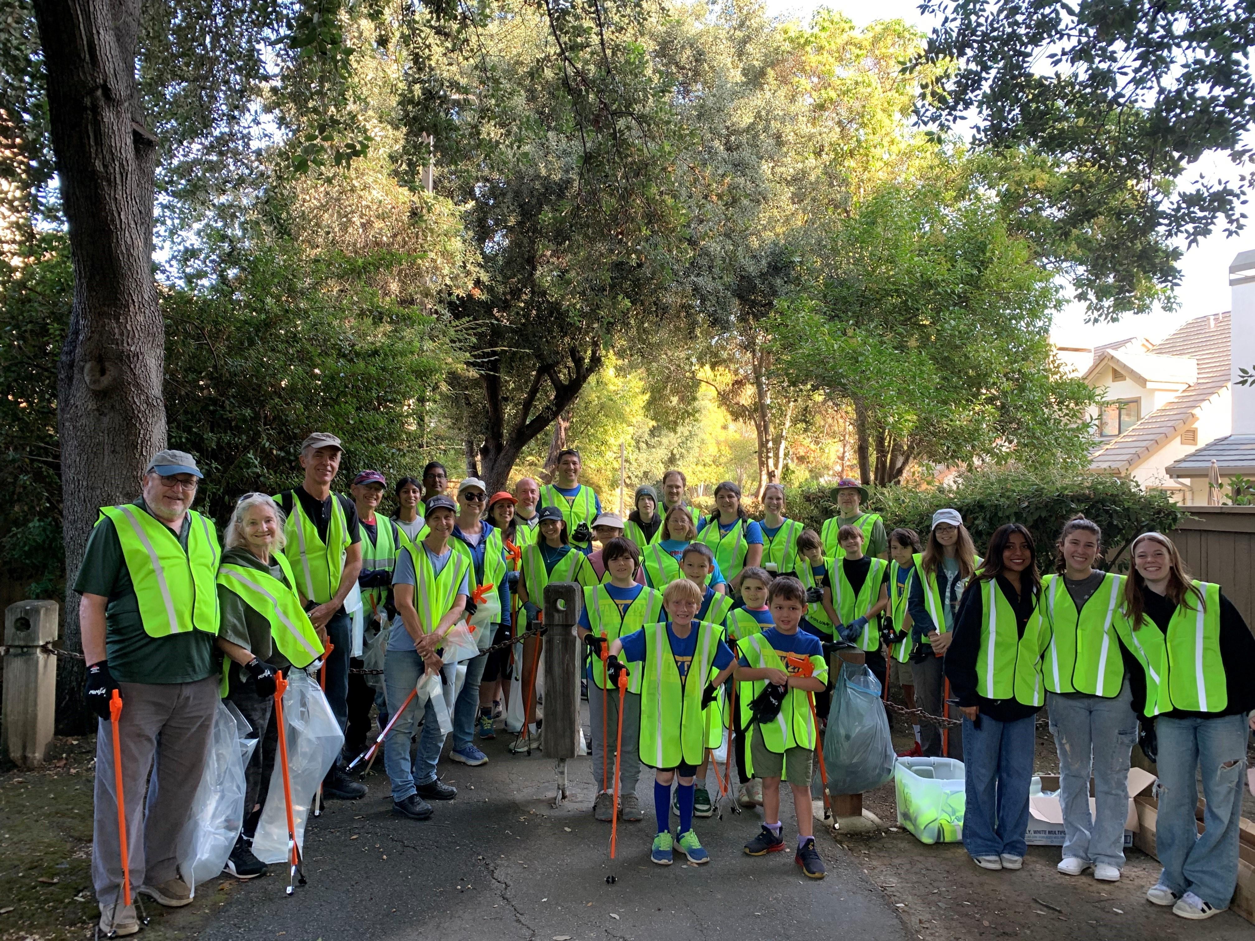 A group of volunteers in yellow vests stand at a cleanup site. A few are holding orange trash grabbers and trash bags.