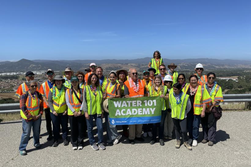 A group of adults holding a banner that reads "Water 101 Academy" standing on the road atop Anderson Dam with a view of South San Jose in the background.